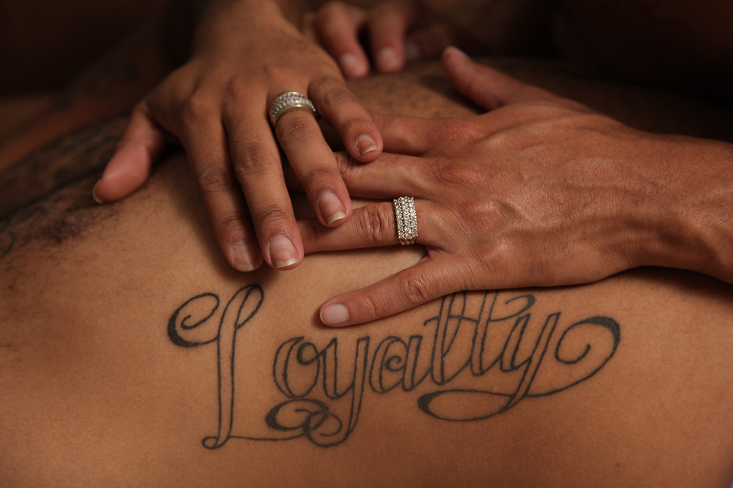 Photograph "Loyalty" by Dawn Hartman. A man's hands on another's back with the tattoo reading "Loyalty" across the spine and shoulder blades. Visit LGBTintimacyproject.org