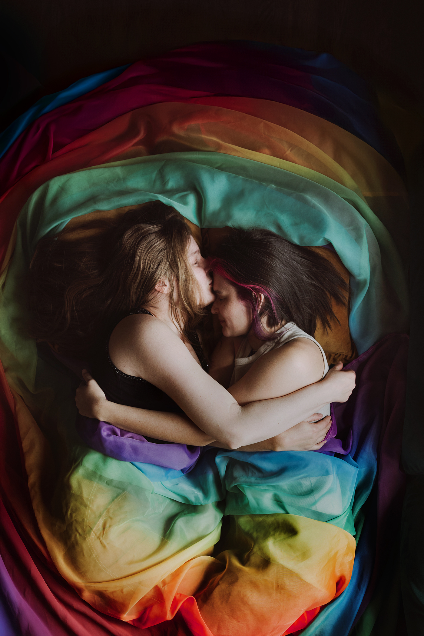 Image 'Over the Rainbow' by Dawn Hartman. Photograph of two people embracing, laying down wrapped only in a rainbow curtain. We wanted to use the rainbow curtain to represent being unapologetically queer