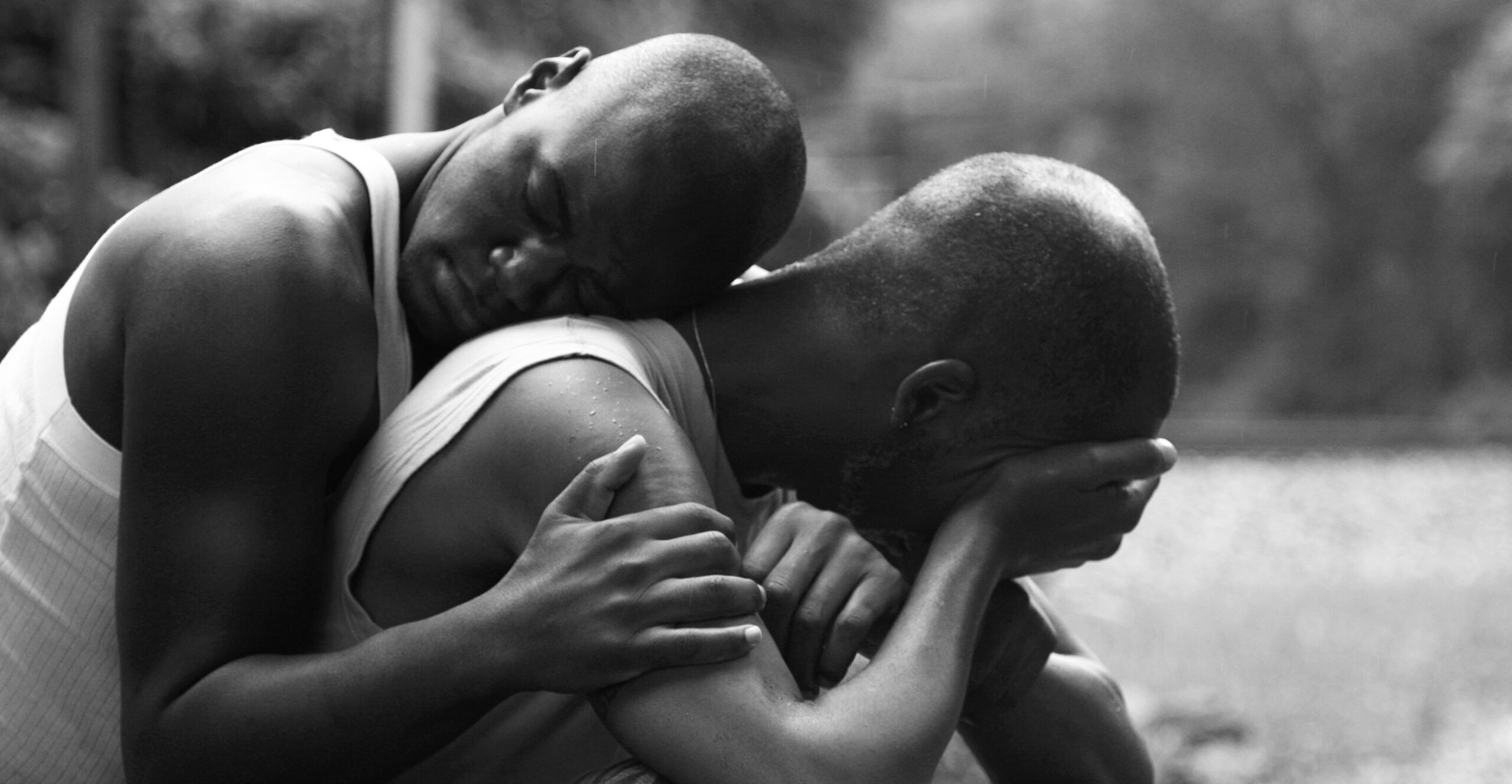 Photograph "Comfort and Strength" by Dawn Hartman. Two people outdoors share a moment: one sits on the ground curled up, and turns away with hand over eyes, his body racked with emotion. The other calmly holds them empathetically. Gently resting head upon their back, they listen silently with eyes closed.